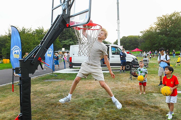 Eine Junge springt an einem Basketballkorb in die Höhe. Andere Kinder jubeln ihm dabei zu.