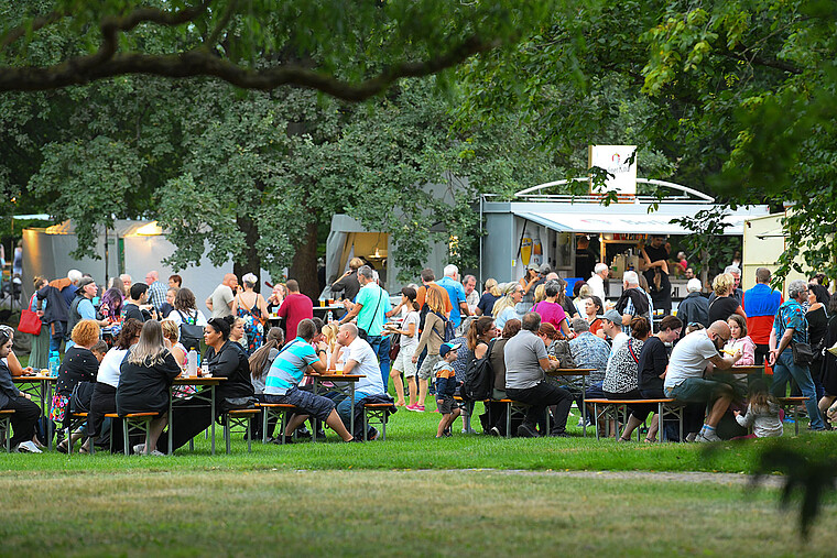 Zu sehen sind Gäste, die in einem Park an Tischen sitzen und essen und trinken. Im Hintergrund sind Verkaufsstände zu sehen. 