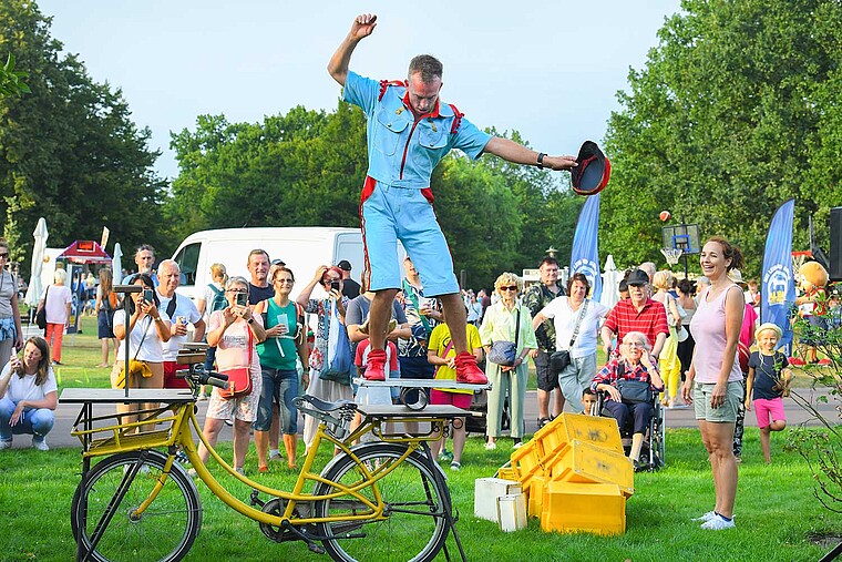 Ein Akrobatik führt auf einem Fahrrad Kunststücke vor. Er tritt im Park auf, die Zuschauer jubeln ihm zu. 