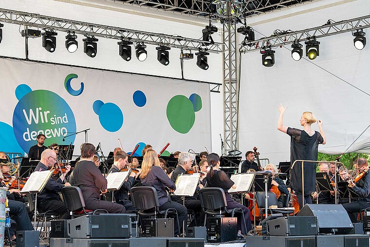 Eine Dirigentin steht auf einer Bühne vor einem Orchester. Im Hintergrund ist das Logo von degewo auf Leinwand zu sehen.