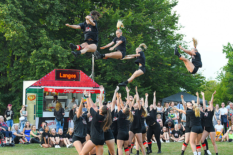 Eine große Gruppe von Cheerleadern zeigt auf einem Fest im Park eine eindrucksvolle Show. Mehrere Mitglieder werden gerade in die Luft geworfen.