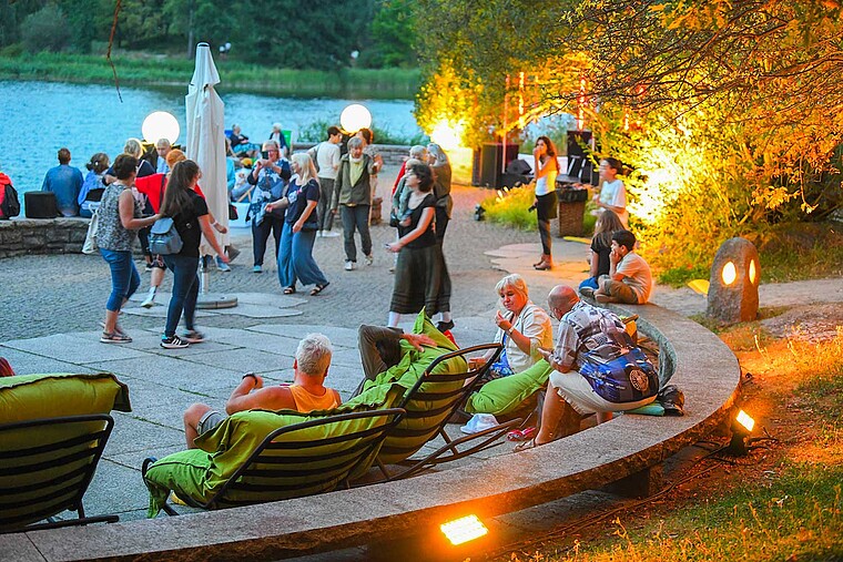 Menschen sitzen an einem See, die Sonne geht unter, die Beleuchtung geht an. Sie unterhalten sich, essen und trinken und genießen die Atmosphäre.