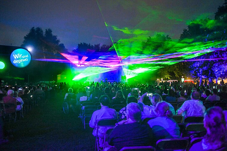 Man sieht eine Lasershow vor großer Bühne. Auf einer großen Rasenfläche davor sitzen oder stehen Menschen und schauen gespannt dabei zu. 